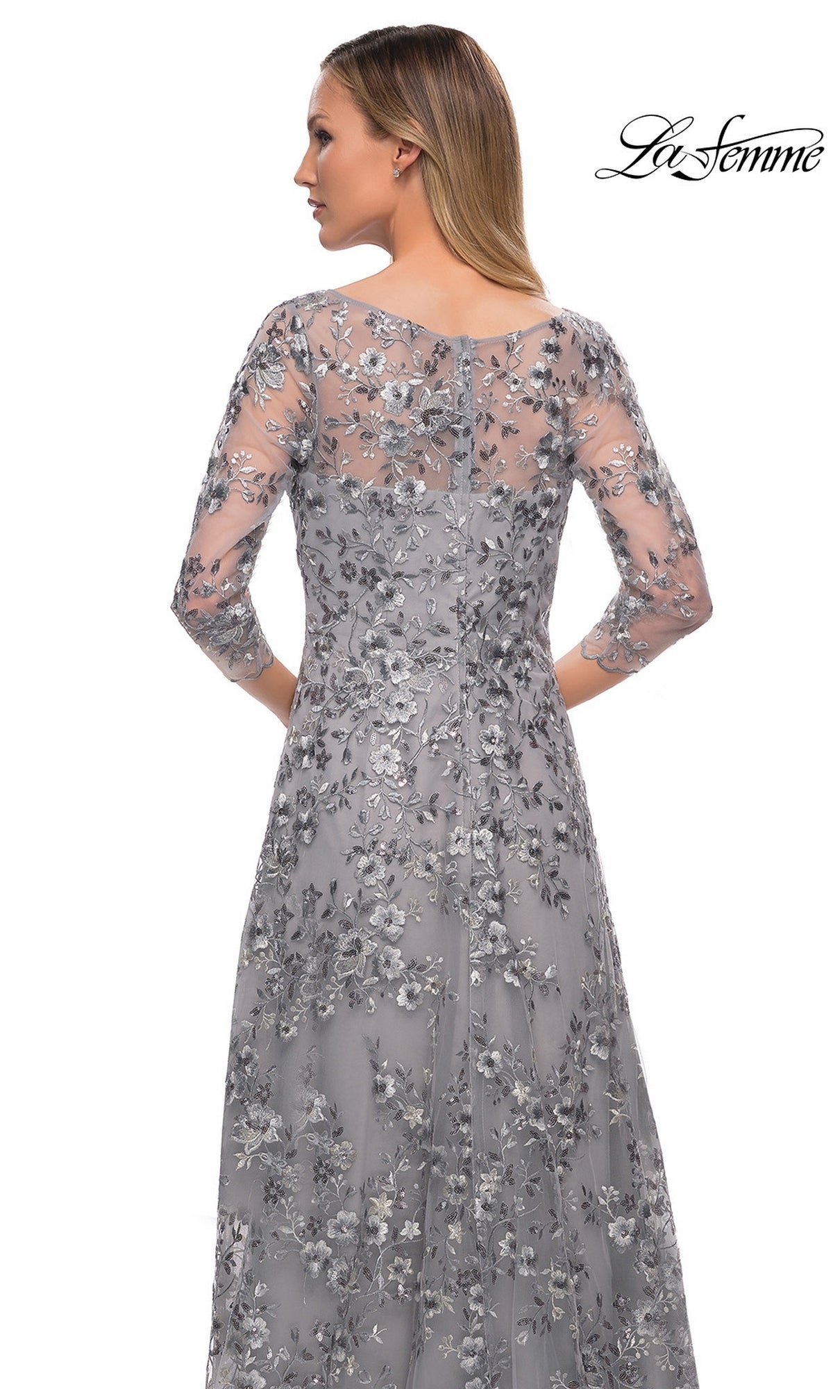 Woman in a silver Long Formal Dress: La Femme 29903 with metallic floral lace, sheer sleeves, and back panel, facing away. “La Femme” logo at upper right.